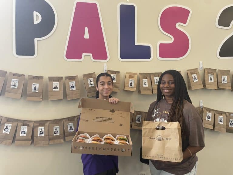 Two NVEEE interns holding donated snacks and a gift bag in front of the PALS banner