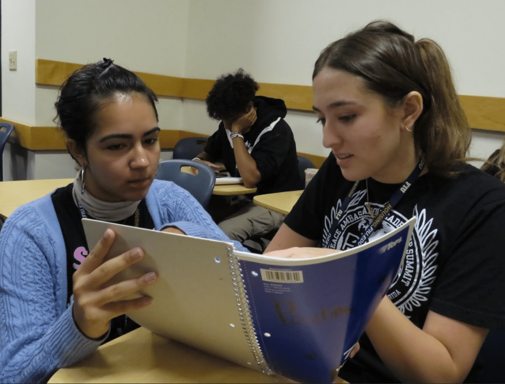 Two girls talking about a notebook they are holding.