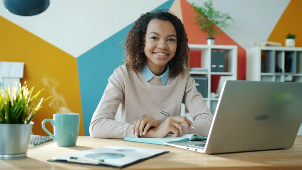 Smiling college student at a home desk with a laptop and coffee, ready to start her shift